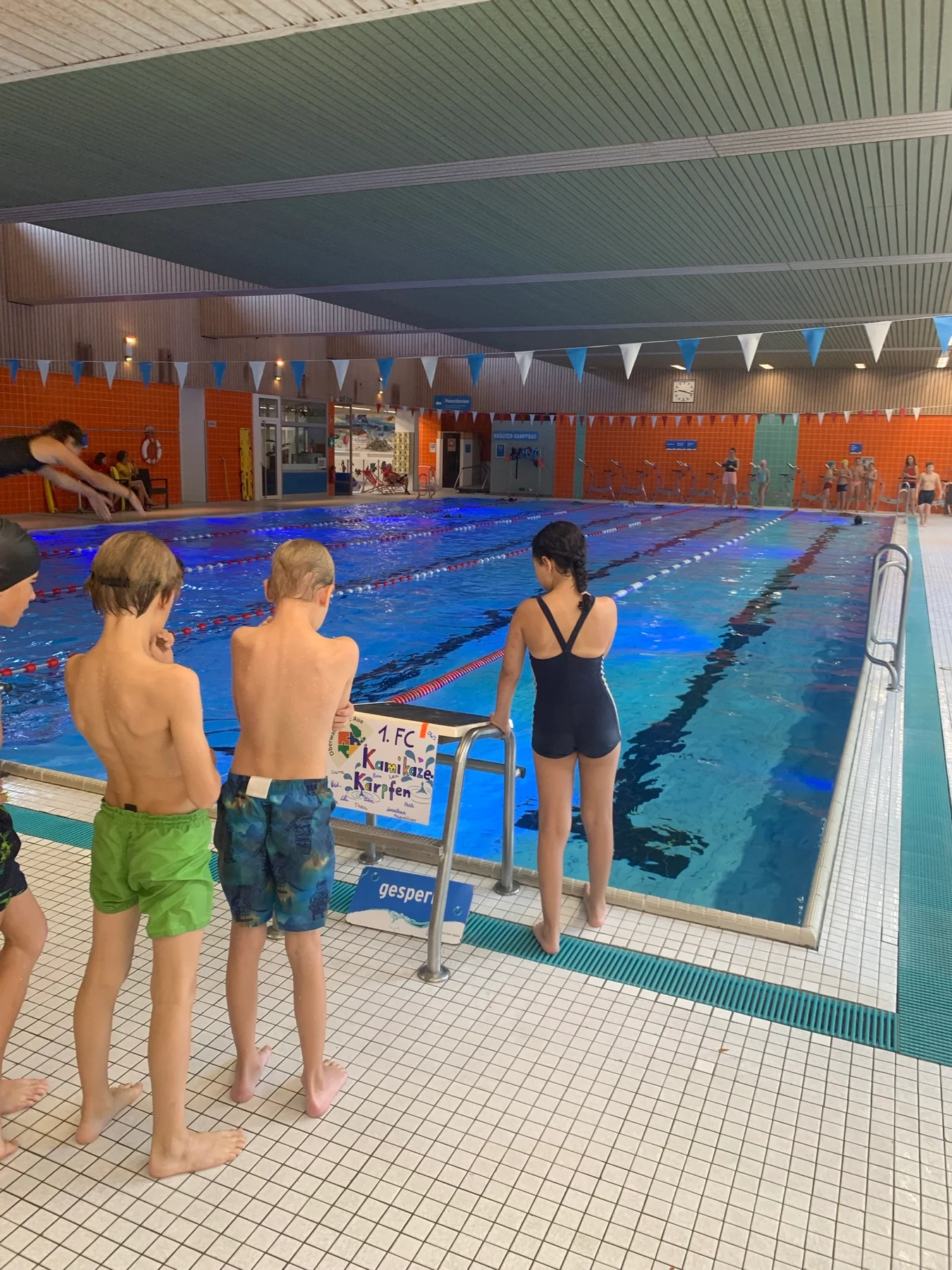 Kinder am Beckenrand im Weiherhofbad; am Startblock hängt das Plakat des Teams 1. FC Kamikaze-Karpfen.